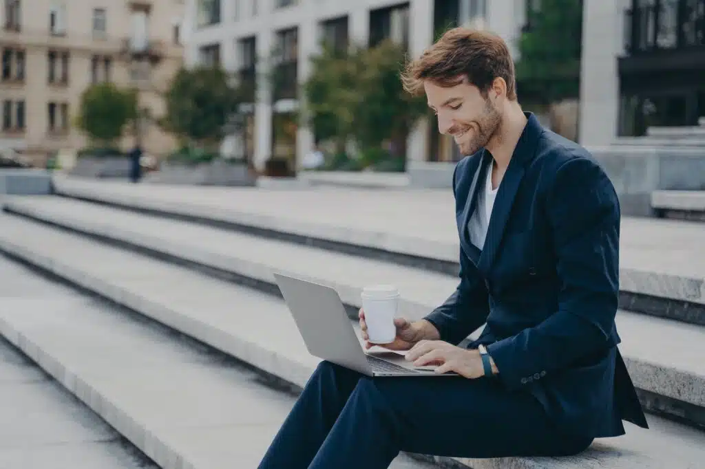 Young satisfied businessman in stylish suit sitting on stairs outside with laptop and coffee to go